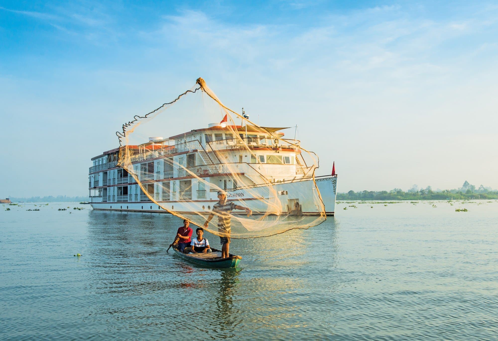 A fisherman on a small watercraft casts a net into the Mekong River with The Jahan riverboat in the background
