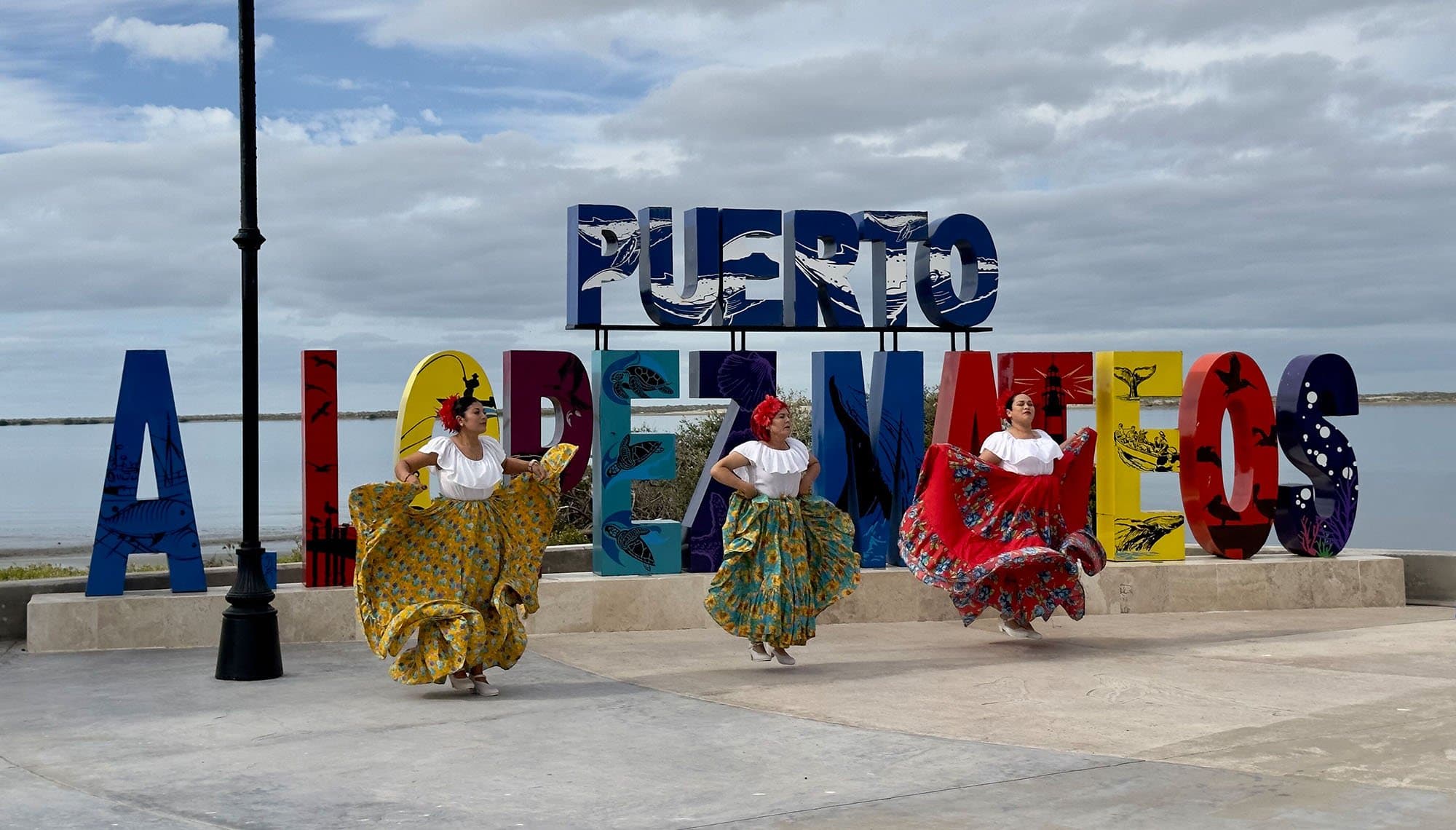three dancers in colorful costumes in front of a colorful sign that says Puerto a Lopez Mateos