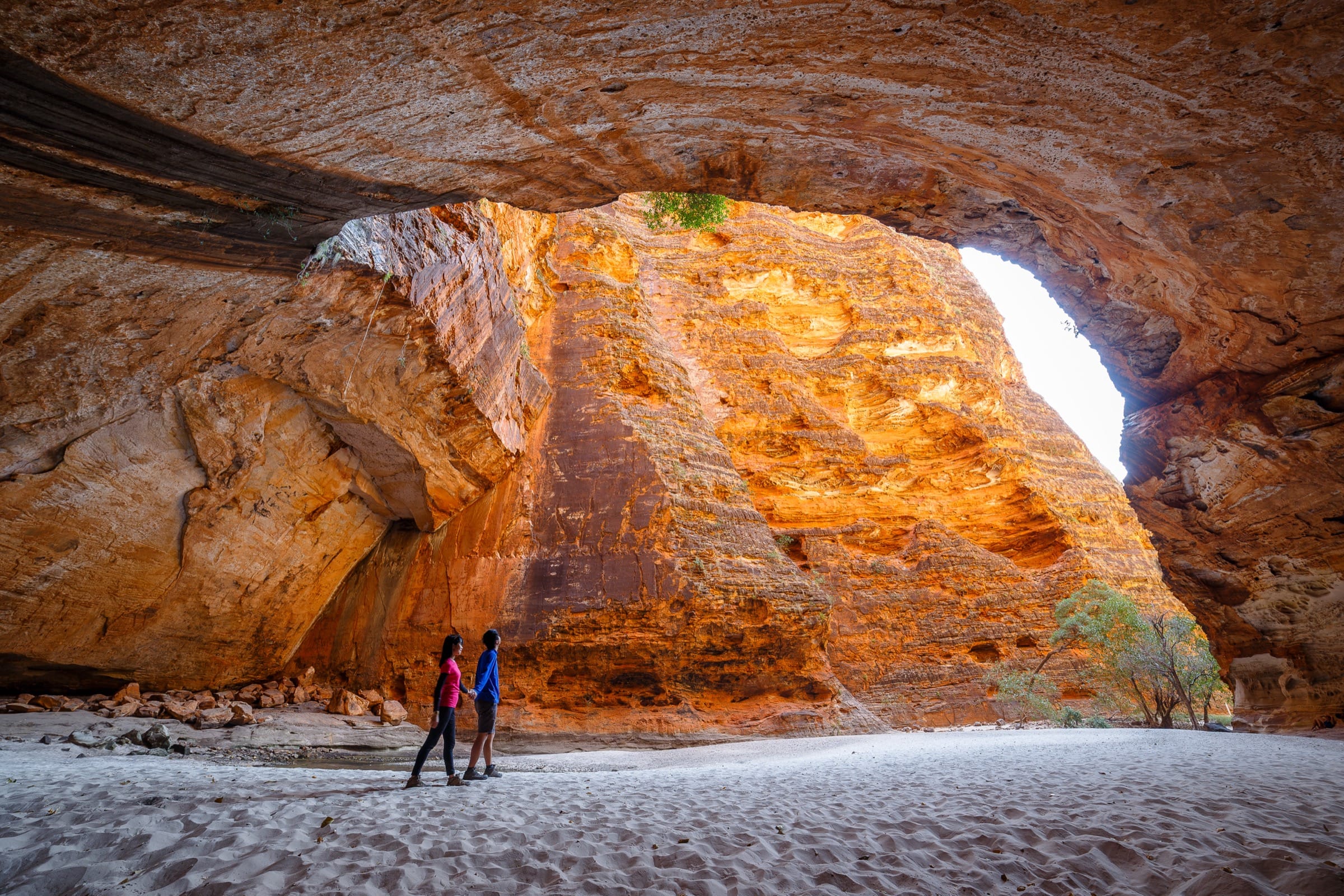 A couple of people holding hands pass through a passage of pure rock, treading soft white sand