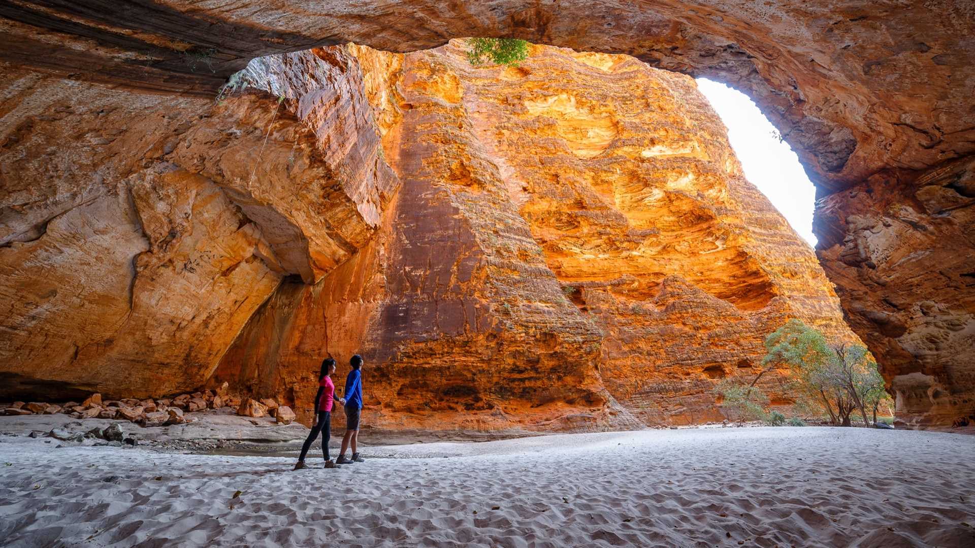 A couple of people holding hands pass through a passage of pure rock, treading soft white sand