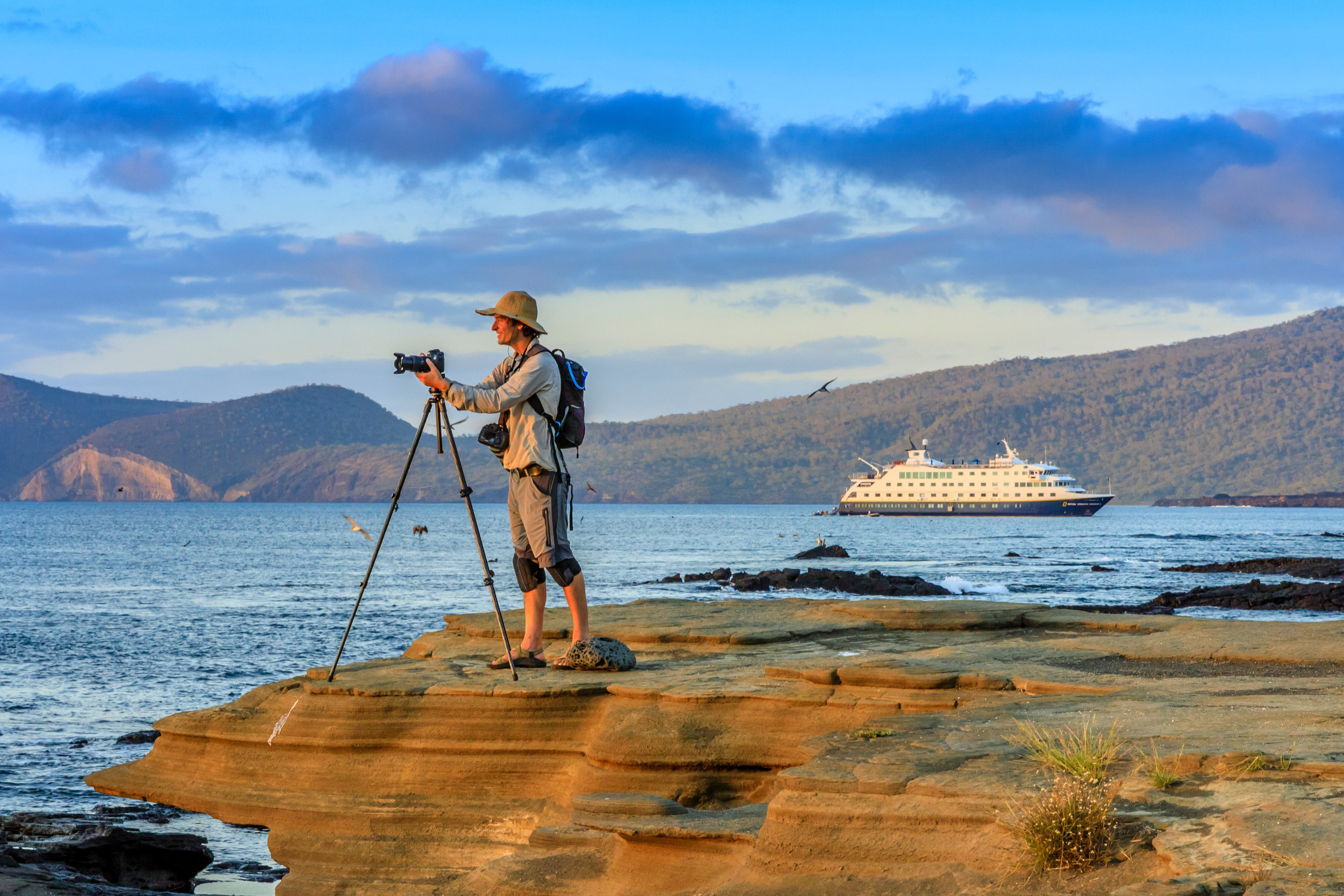 A photographer takes a photo at sunset on the rocky shores of Santiago Island, Galápagos, with the ship National Geographic Endeavour II in the background.