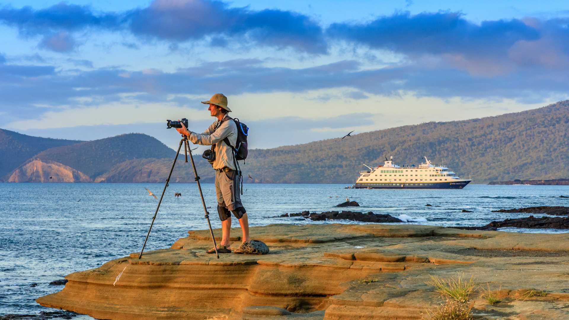 A photographer takes a photo at sunset on the rocky shores of Santiago Island, Galápagos, with the ship National Geographic Endeavour II in the background.