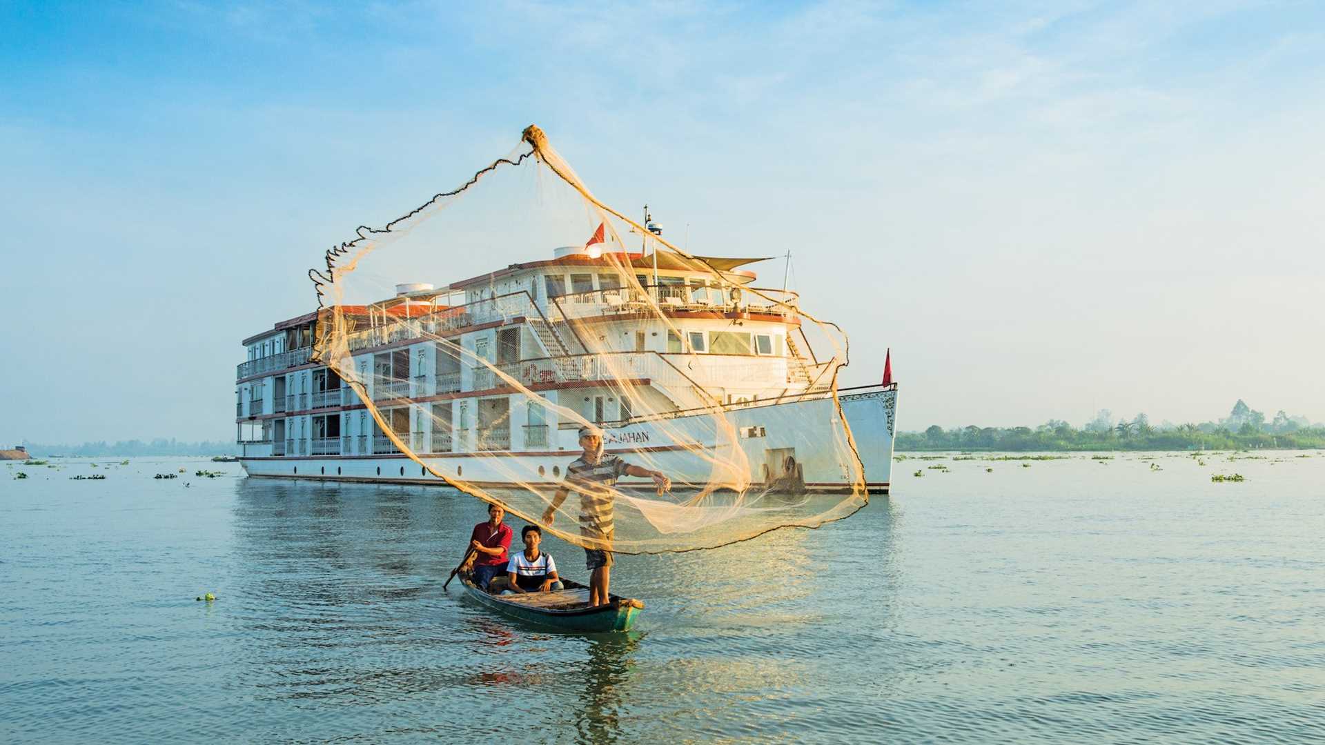 A fisherman on a small watercraft casts a net into the Mekong River with The Jahan riverboat in the background