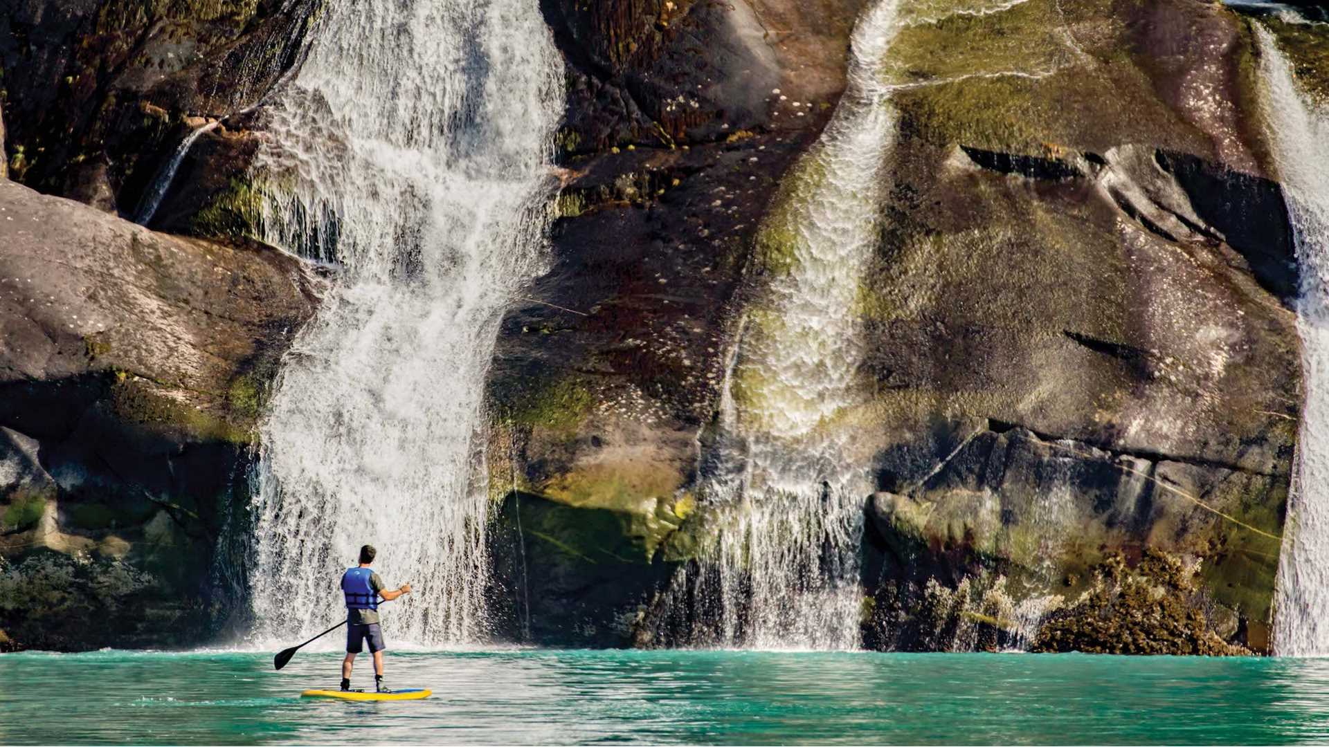 A solo paddleboarder stops in front of a waterfall