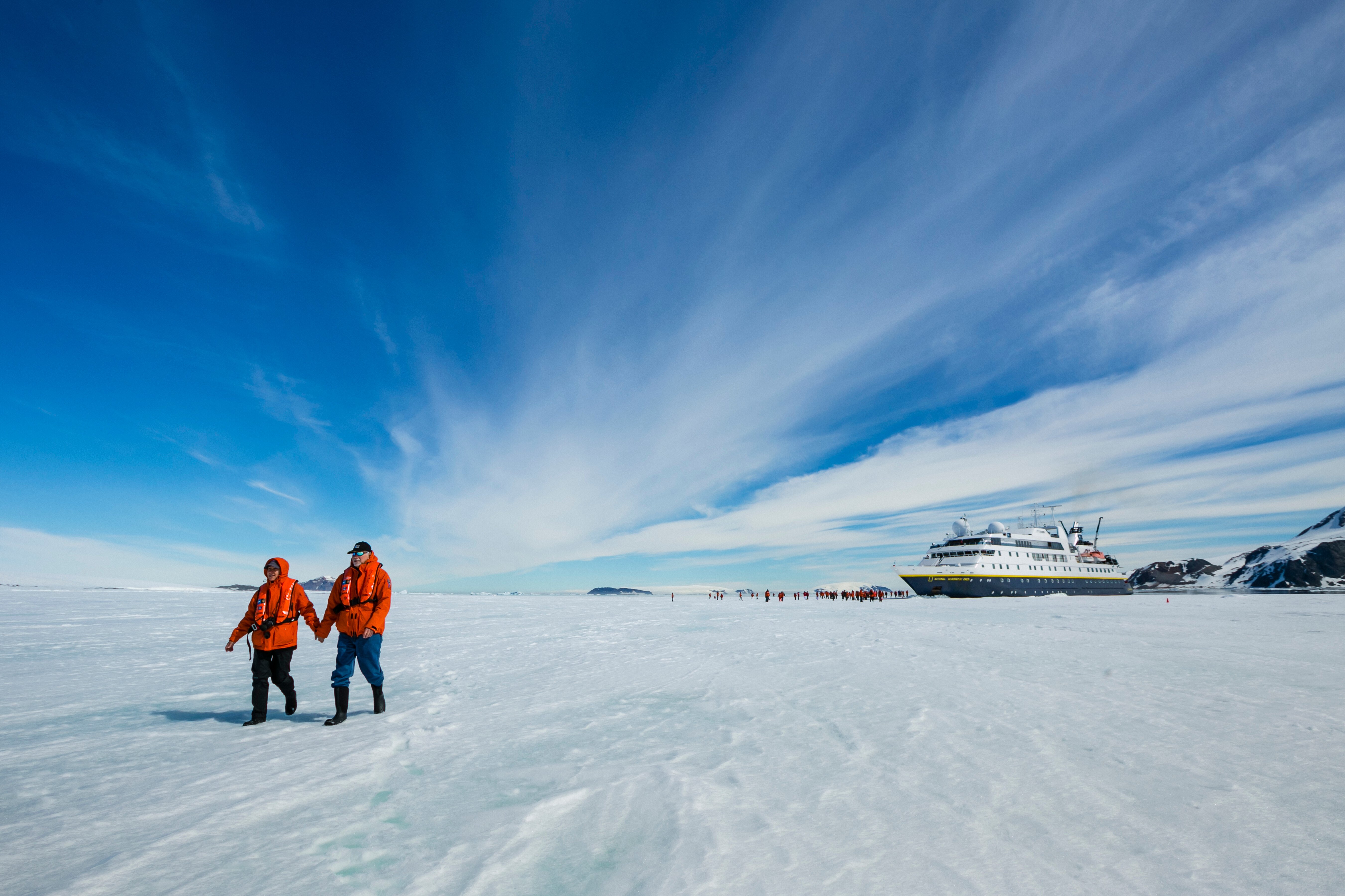 Two guests hold hands as they walk on pack ice with the National Geographic Orion in the distance.