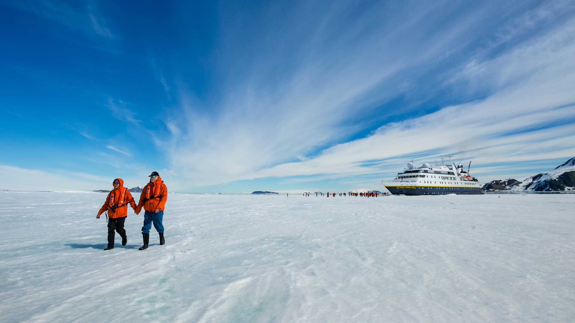 Two guests hold hands as they walk on pack ice with the National Geographic Orion in the distance.