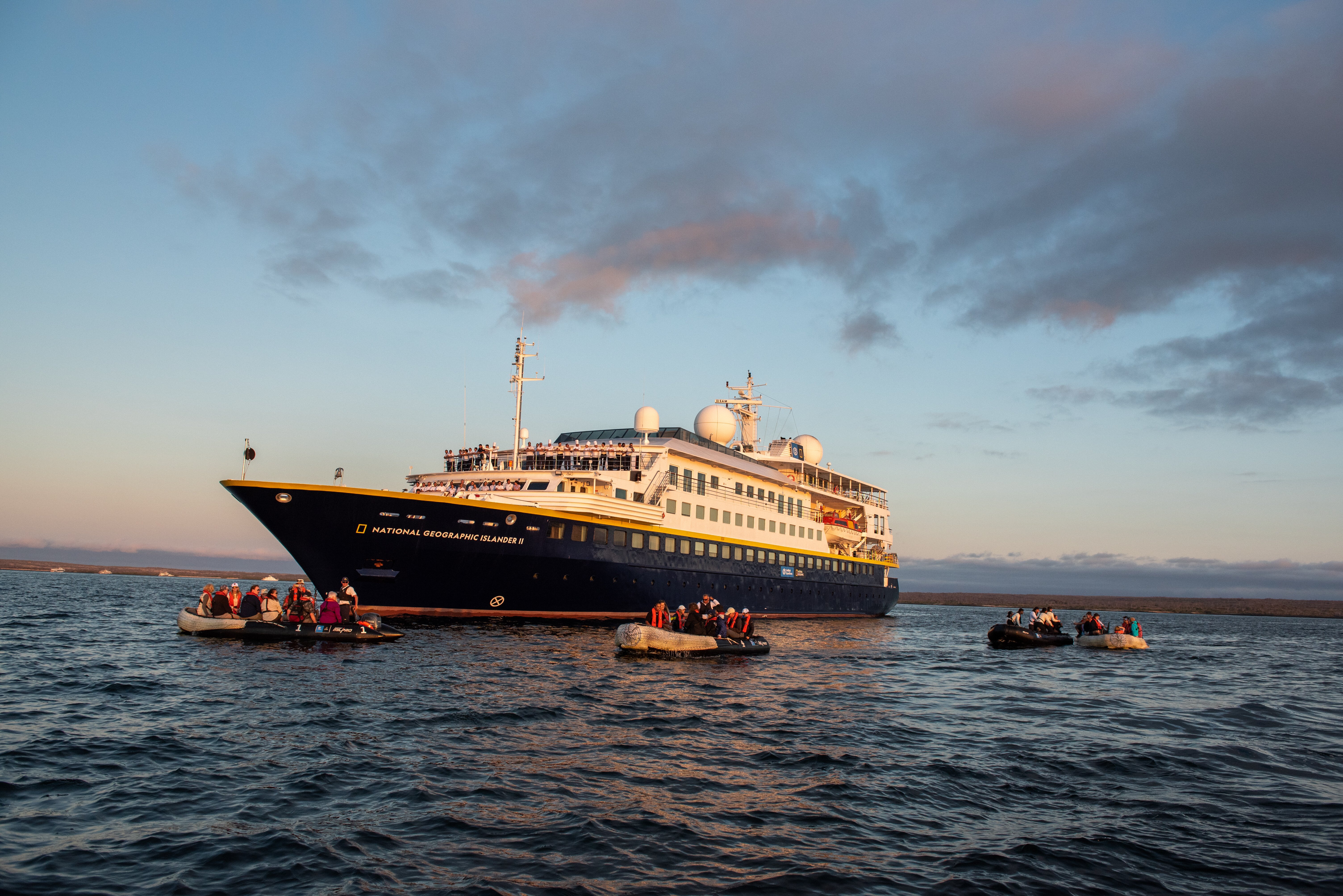 The ship National Geographic Islander II anchored at sunset with five Zodiacs parked in front.