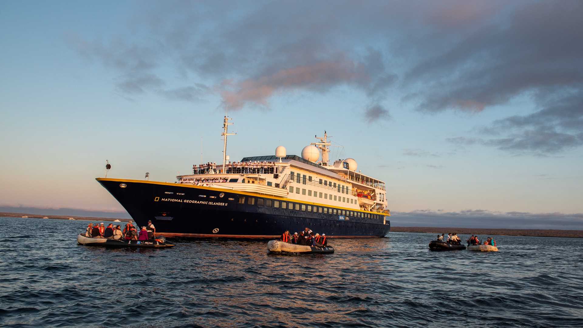 The ship National Geographic Islander II anchored at sunset with five Zodiacs parked in front.