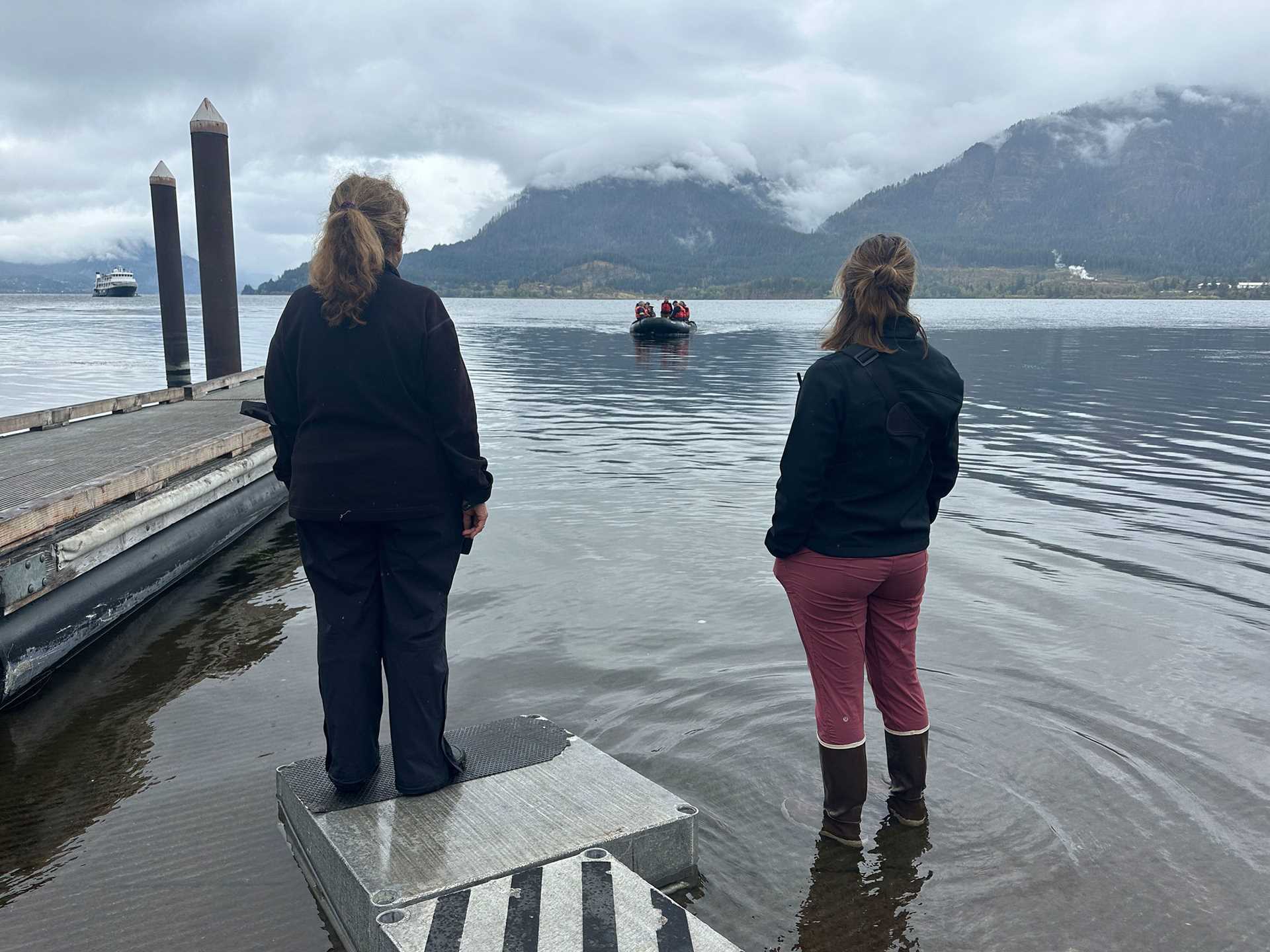 two people stand in the water watching a zodiac craft coming in