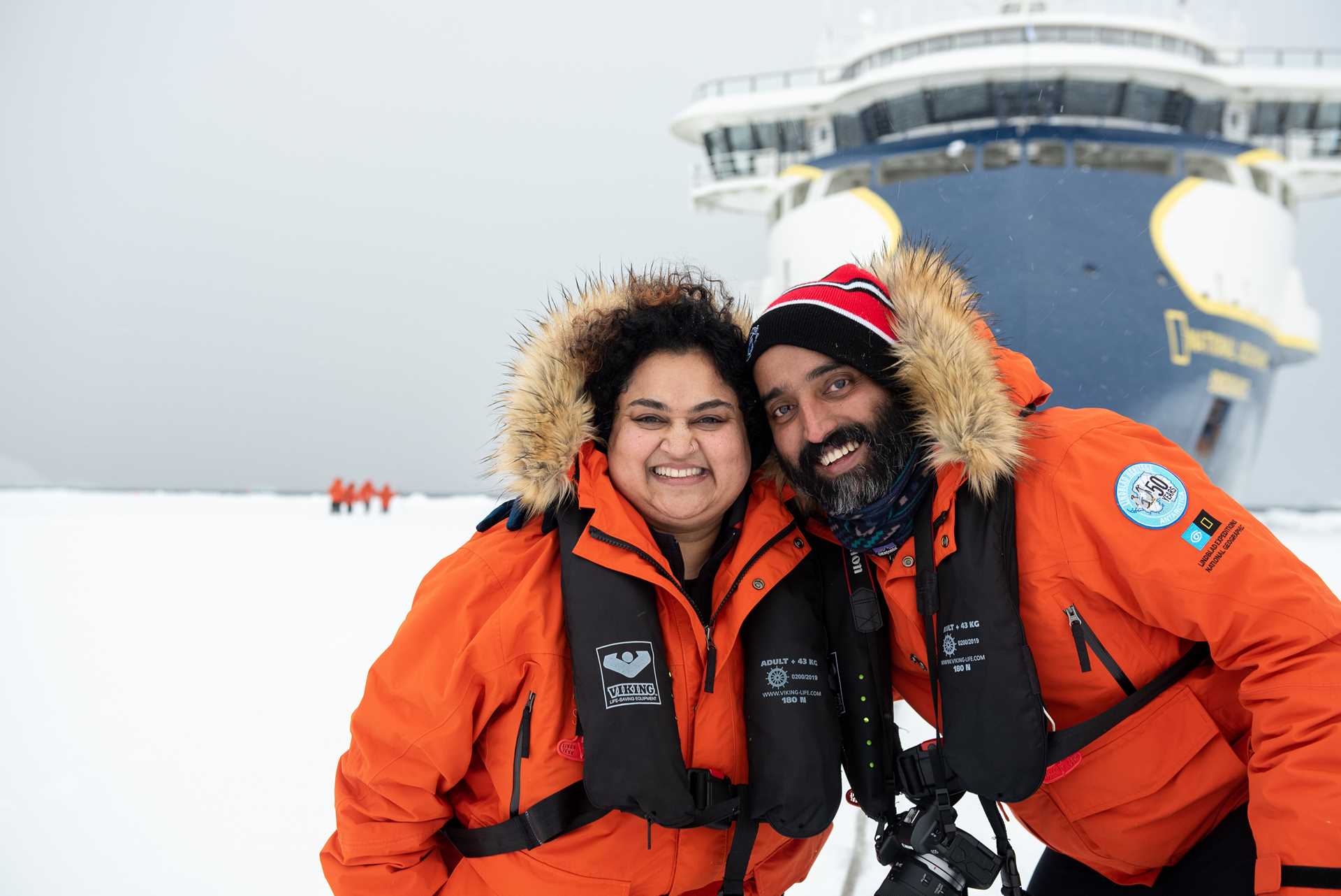 Two guests pose for a photo standing on fast ice in front of the National Geographic Endurance.